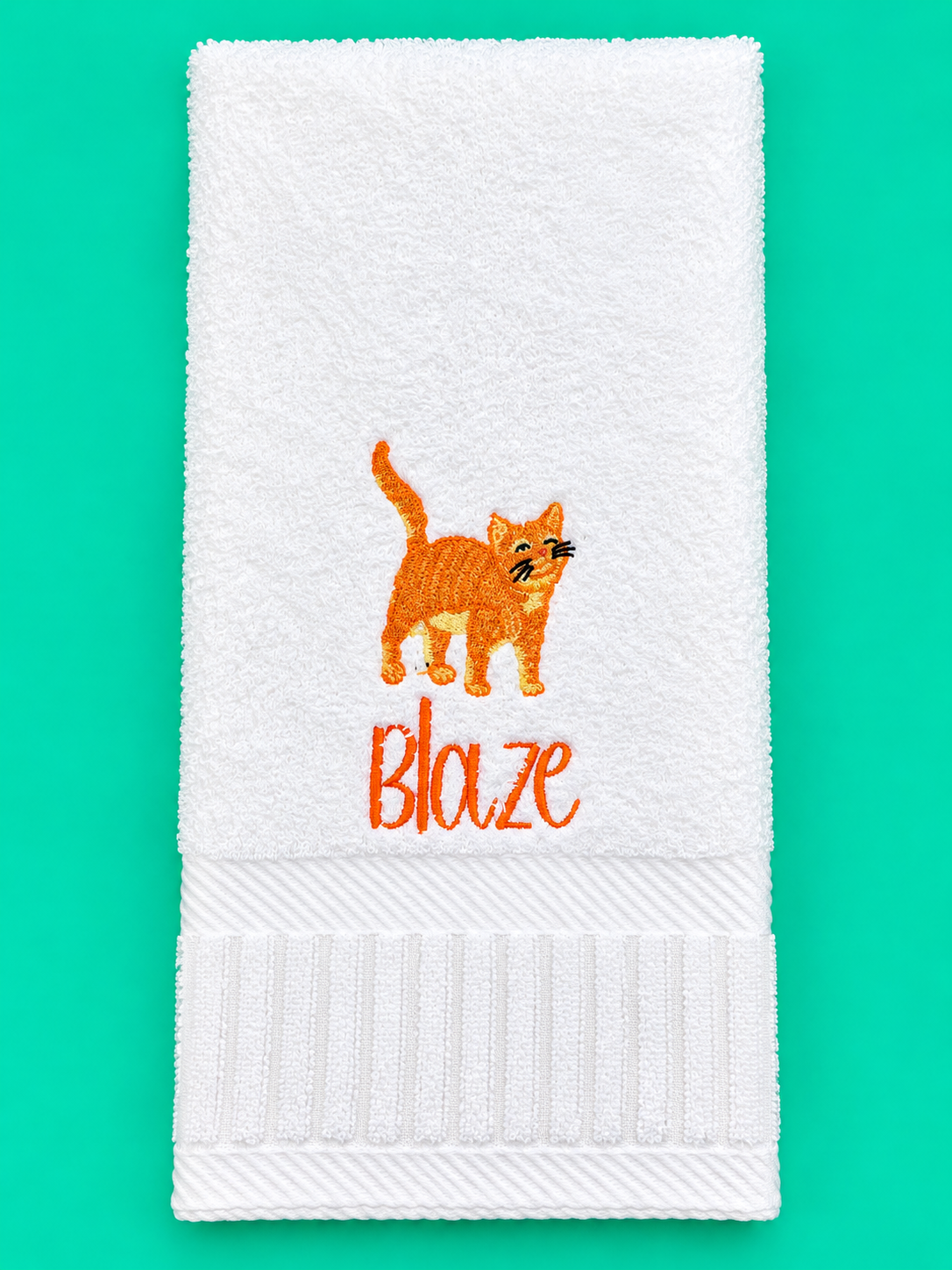 White hand towel embroidered with an orange cat and Blaze on turquoise green