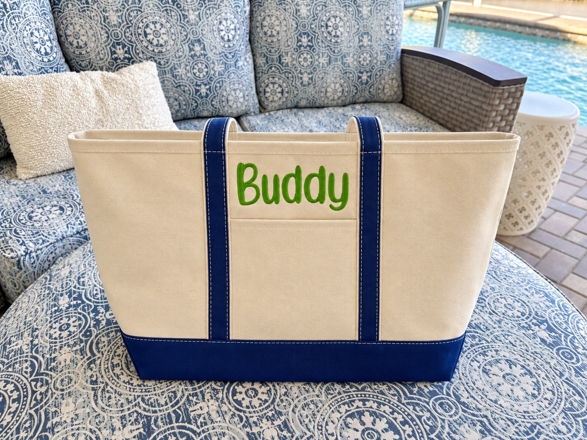 Cream canvas tote with navy trim and green Buddy embroidery on a coastal patio sofa