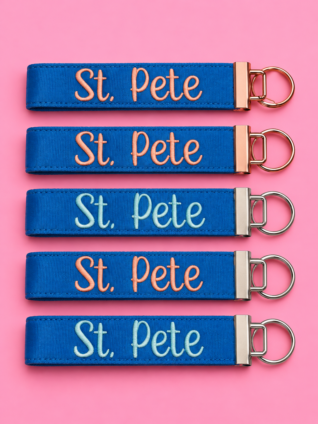 Five blue St. Pete embroidered wristlet keychains on a pink background